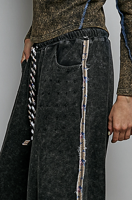 Close-up of a person wearing dark charcoal corduroy wide-leg pants with a decorative multicolor frayed stripe down the side and a braided drawstring at the elastic waistband.