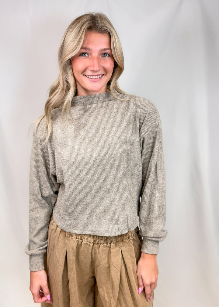 Woman wearing beige mock neck knit top with brown pants on plain background