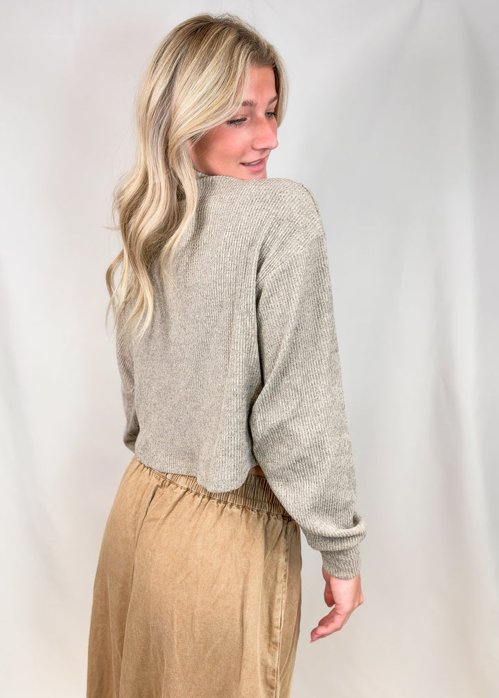 A woman with blonde hair is shown from the back wearing a light beige ribbed knit cropped sweater and a tan skirt, standing against a white fabric backdrop.