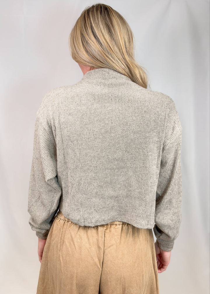 A woman viewed from behind wearing a light gray ribbed knit cropped sweater and tan pants, standing against a white studio backdrop.