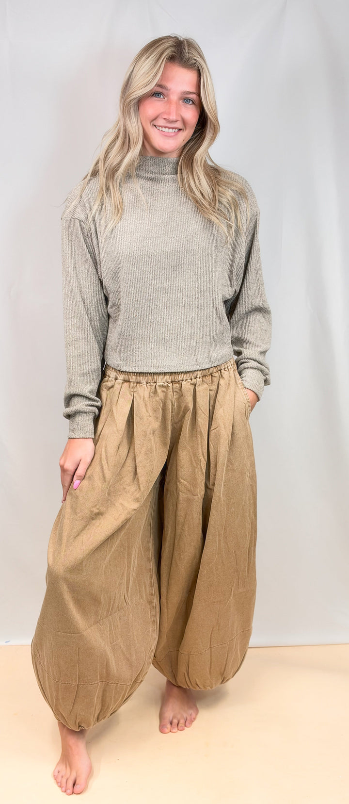 A woman stands barefoot against a light neutral studio backdrop, wearing a light gray turtleneck sweater and loose, baggy tan pants with gathered hems and side pockets.