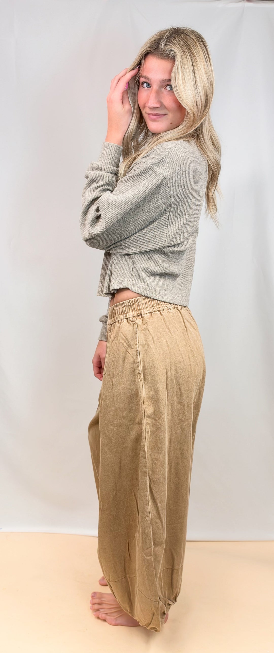 A woman stands barefoot against a white studio backdrop, wearing a light gray cropped knit cardigan and loose beige wide-leg pants.