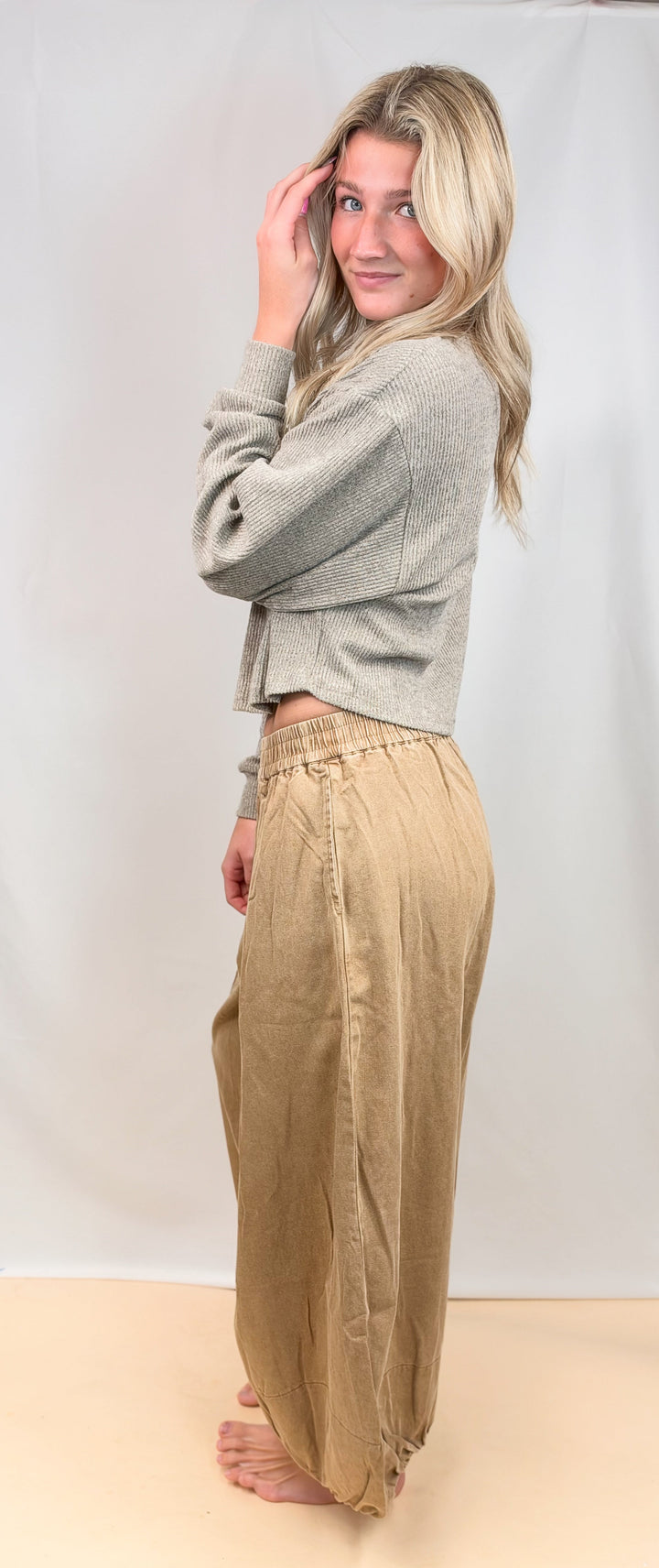 A woman stands barefoot against a white studio backdrop, wearing a light gray cropped knit cardigan and loose beige wide-leg pants.