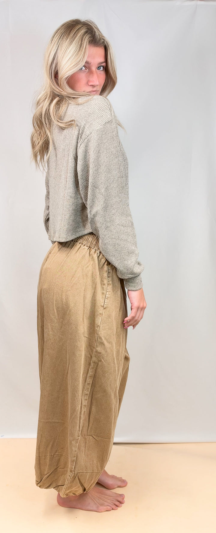 A woman stands barefoot in a studio wearing a light gray cropped sweater and loose tan pants, photographed against a plain light backdrop.