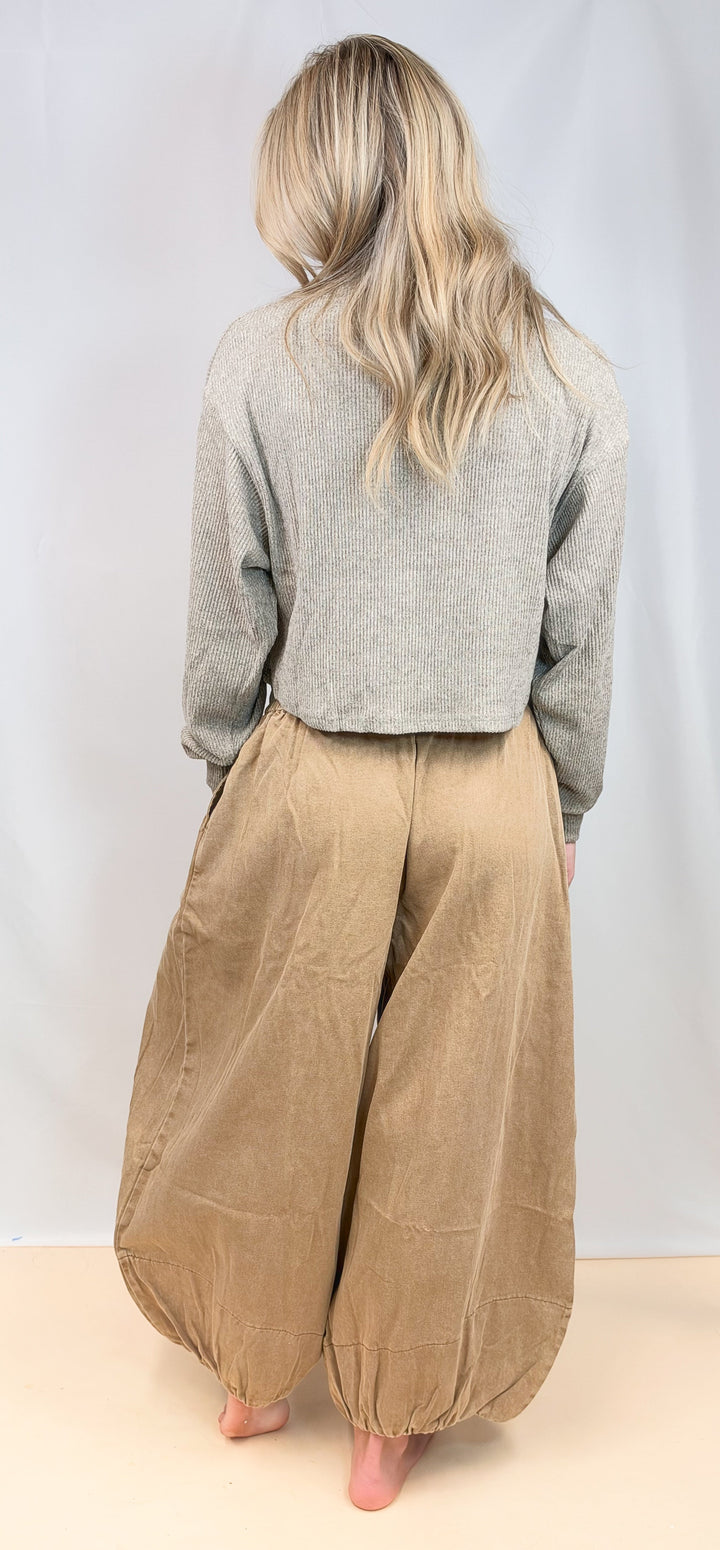 A woman shown from behind wearing a light gray long-sleeve cropped sweater and voluminous tan corduroy harem pants, standing barefoot against a plain light gray studio backdrop.