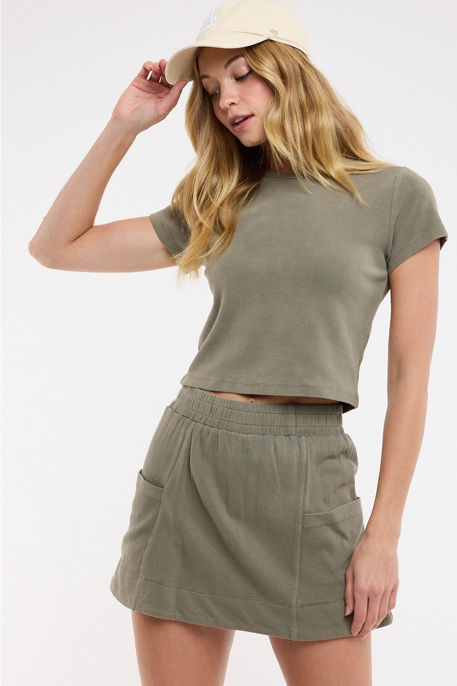 Woman wearing an olive green crop top and skirt set with a beige cap on a white background