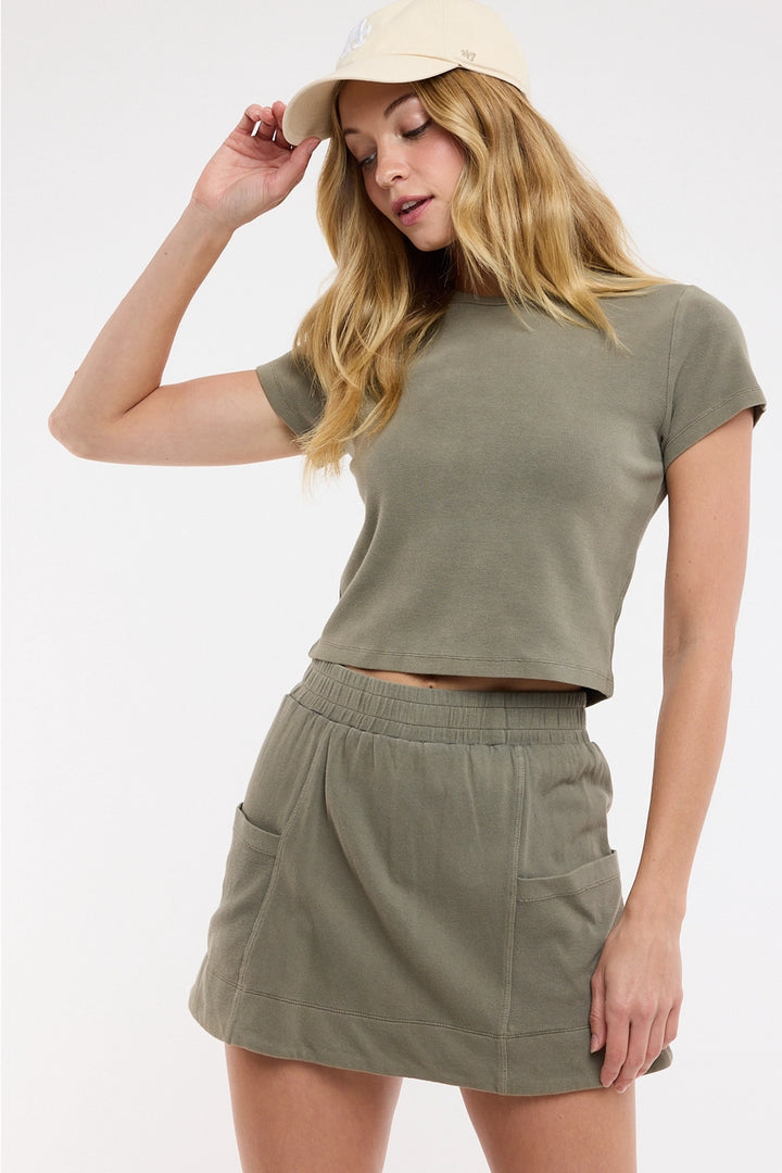 Woman wearing an olive green crop top and skirt set with a beige cap on a white background