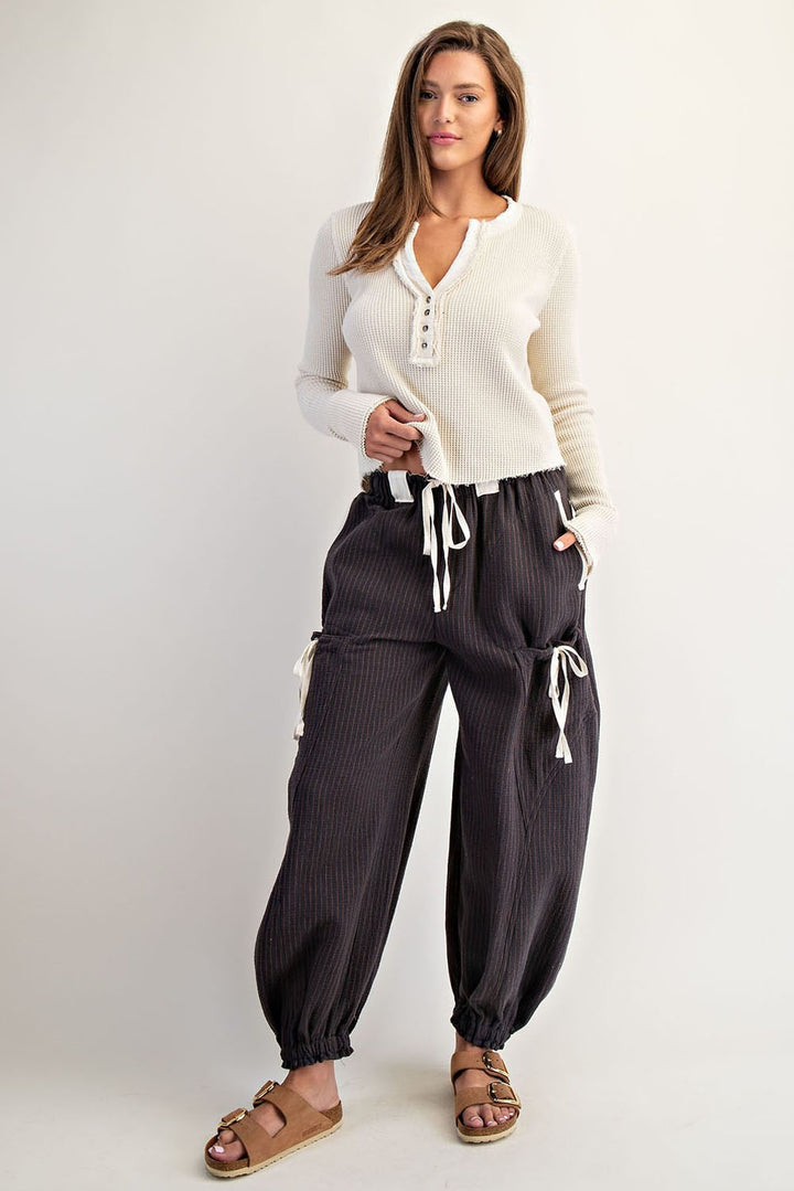A woman modeling a cream ribbed long-sleeve henley top and dark charcoal gray pinstriped harem pants with white drawstrings, paired with brown sandals, against a light gray studio background.