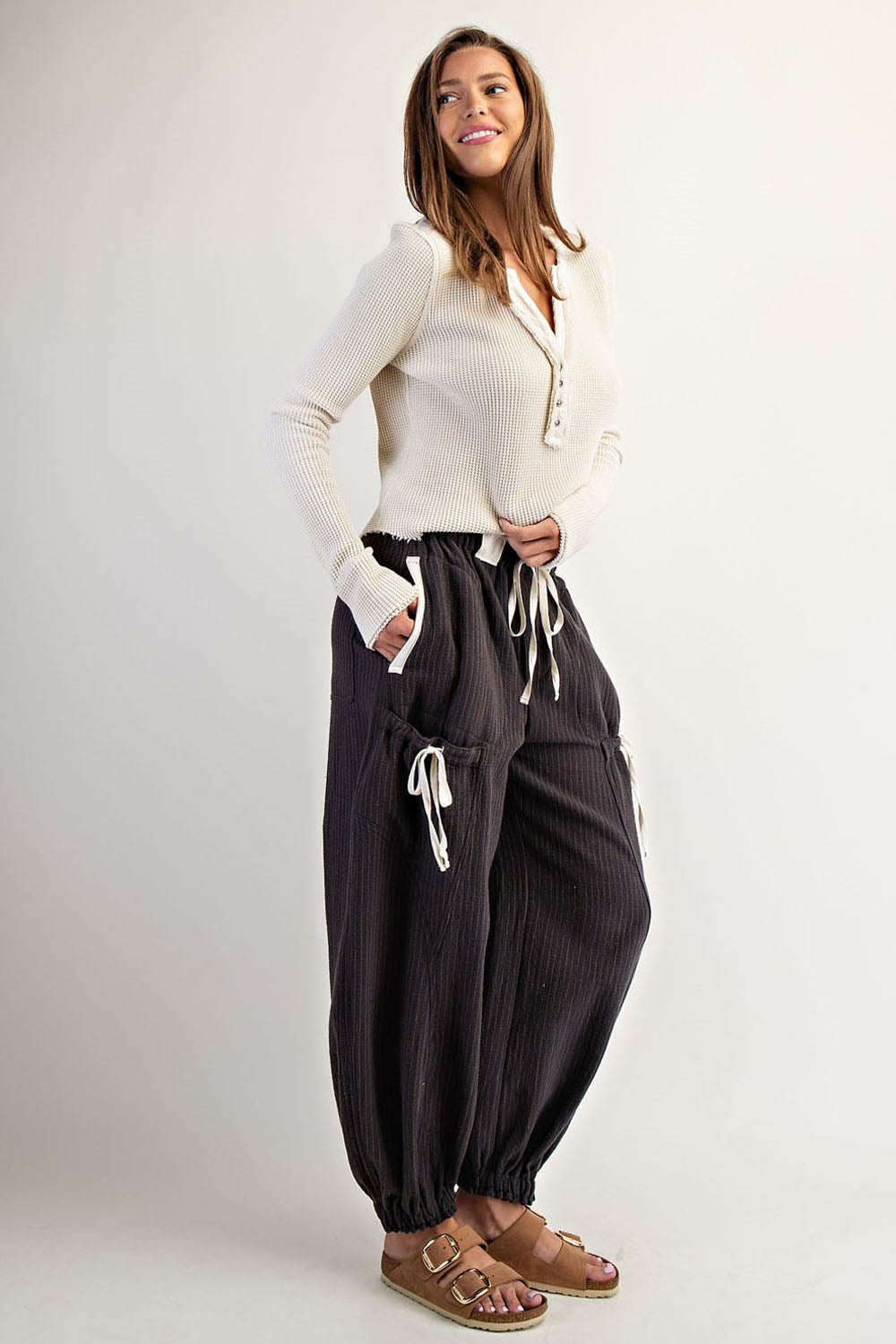 A woman modeling loose dark charcoal pleated harem pants with white drawstrings, paired with a cream ribbed long-sleeve top and brown sandals, against a light gray studio background.
