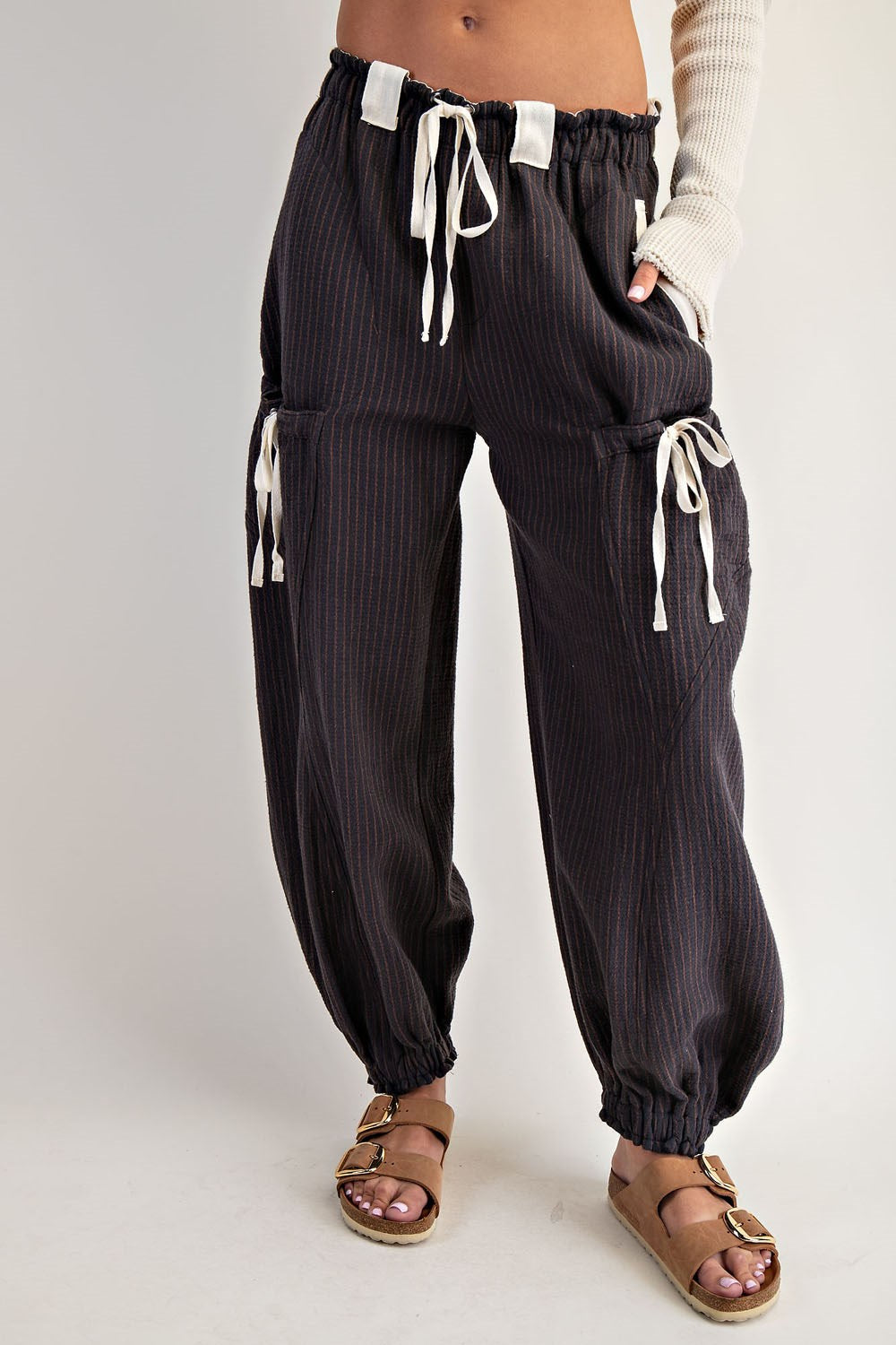 A person wearing loose dark pinstriped harem-style pants with white drawstrings and side tie details, paired with tan sandals and a light long-sleeve top, photographed against a plain light background.