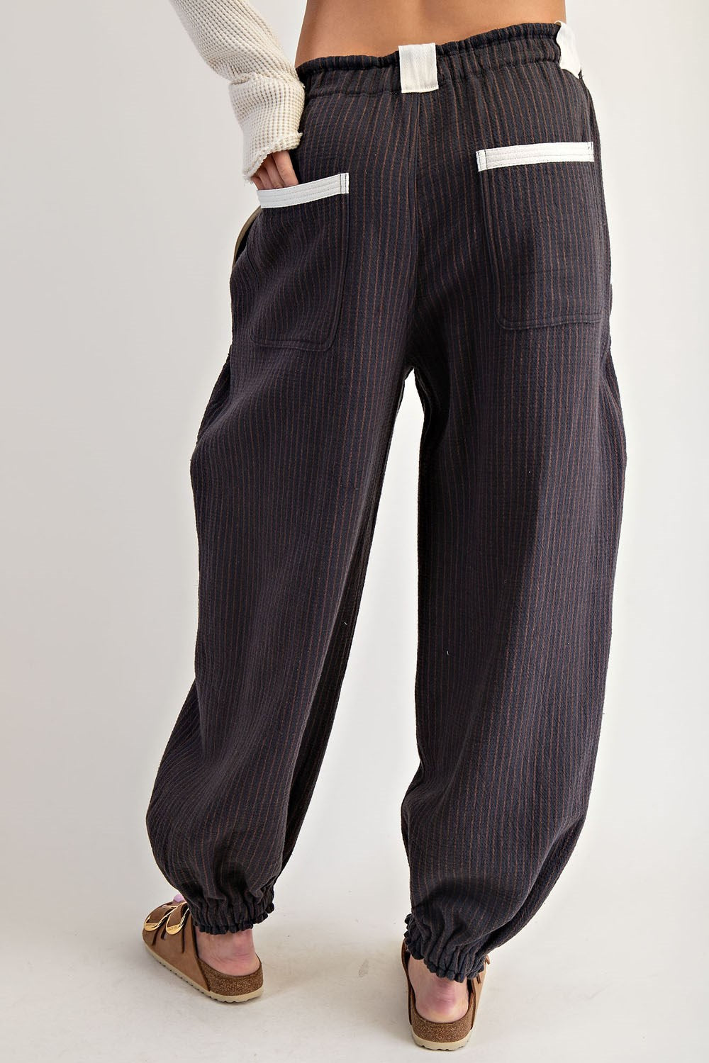 A person shown from the back wearing loose dark charcoal pinstriped pants with white contrast stitching on the pocket edges, a white ribbed knit top, and brown slide sandals, against a light gray studio background.