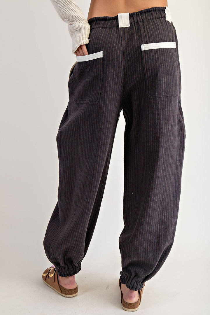 A person shown from the back wearing loose dark charcoal pinstriped pants with white contrast stitching on the pocket edges, a white ribbed knit top, and brown slide sandals, against a light gray studio background.
