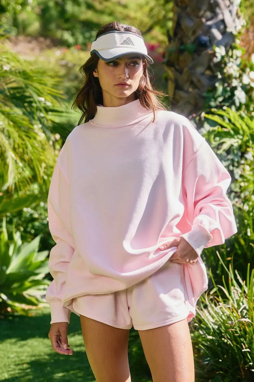 Woman wearing a pink sweatshirt and shorts set with a visor outdoors.