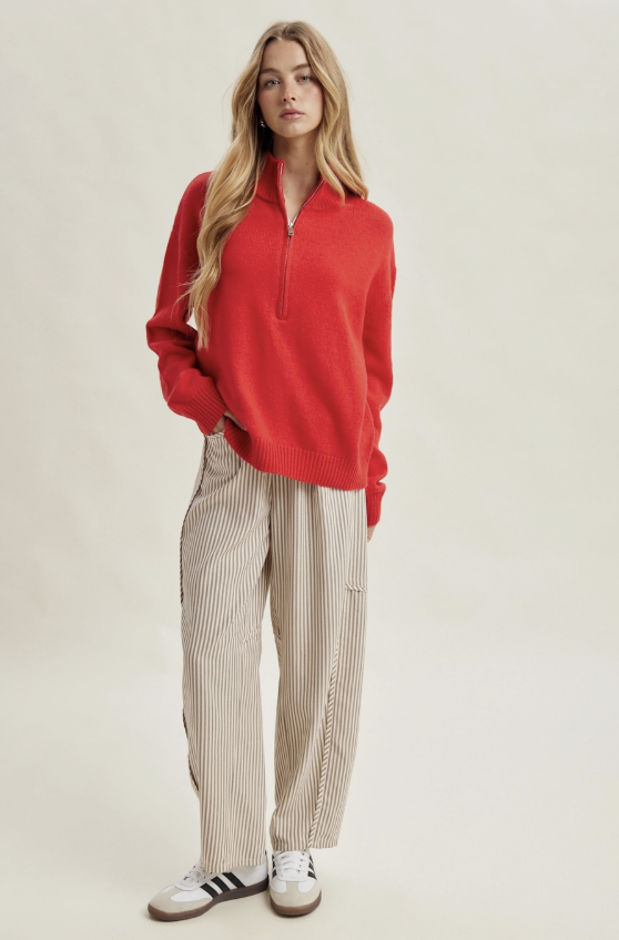 A woman modeling a red half-zip long-sleeve sweater and beige pinstriped trousers with white sneakers, standing against a plain light beige studio background.