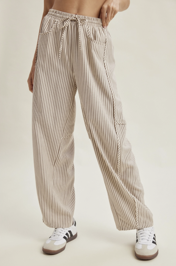 A person wearing loose, wide-leg striped pants in beige and white, paired with white sneakers, shown from the waist down against a plain light beige studio background.