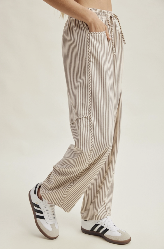 A person wearing loose beige and white vertically striped drawstring pants with side pockets, paired with white low-top sneakers, shown against a plain light beige studio background.