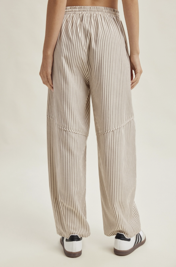 A person shown from the back wearing loose, wide-leg striped pants in beige and white, paired with white sneakers, against a plain light beige studio background.