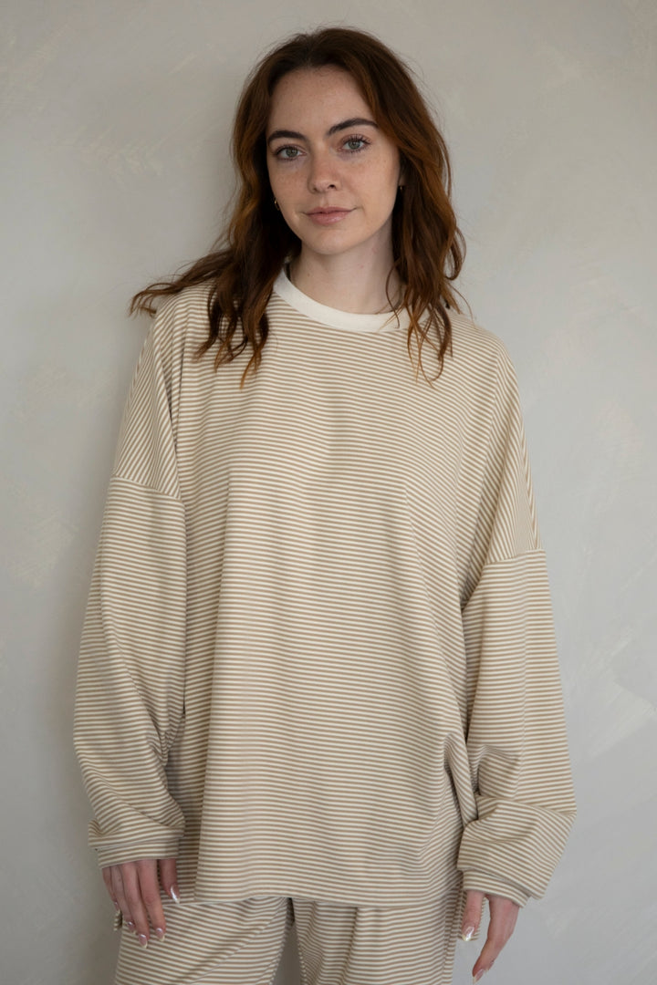 A woman wearing a beige and white horizontally striped long-sleeve crewneck sweatshirt and matching sweatpants, standing against a neutral light gray wall.