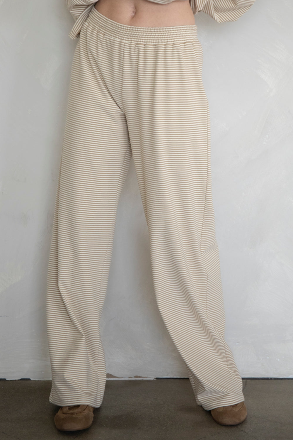 A person wearing loose, wide-leg pants with thin horizontal beige and white stripes, paired with brown shoes, standing against a light textured wall.