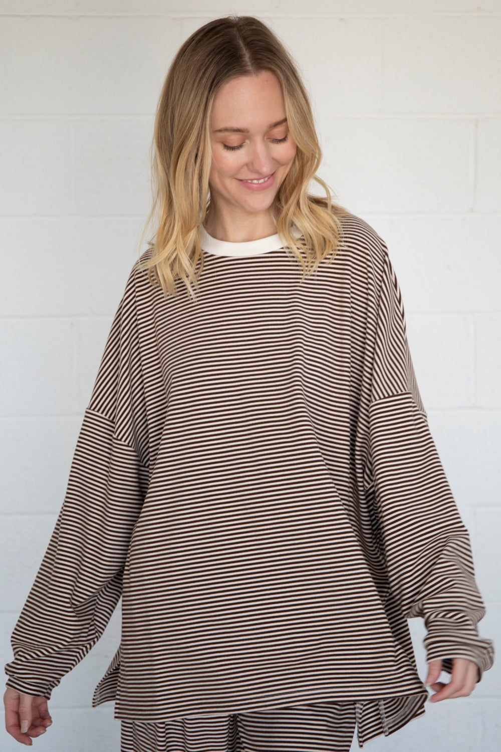 A woman wearing a brown and beige horizontally striped long-sleeve top with a white crew neck, standing against a white brick wall.