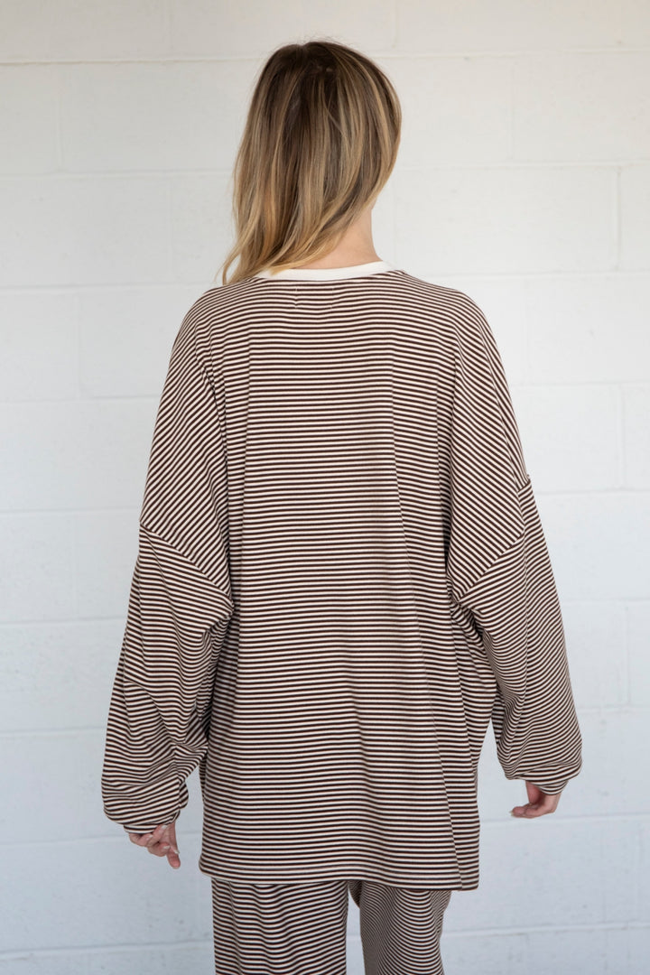 A woman viewed from behind wearing a brown and white horizontally striped long-sleeve top and matching striped pants, standing against a white brick wall.