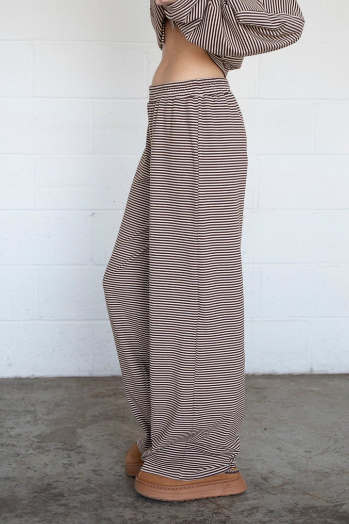 A person shown from the waist down wearing loose, wide-leg striped pants in brown and white, paired with brown platform shoes, standing against a white brick wall.