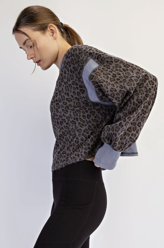 A woman wearing a leopard-print long-sleeve top with light blue contrast panels on the shoulders and sleeves, paired with black leggings, photographed against a plain light gray studio background.