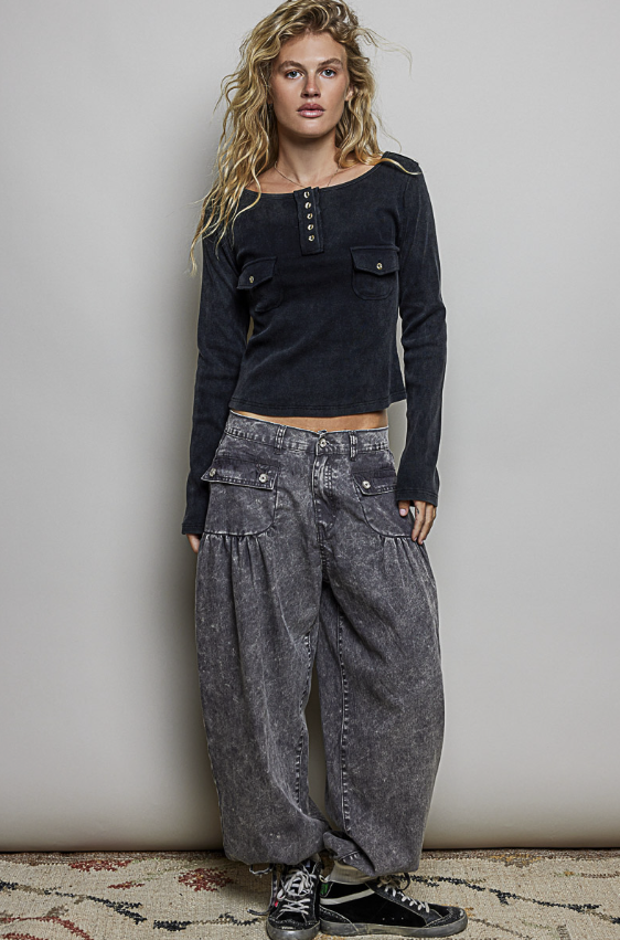 A woman modeling a dark charcoal long-sleeve cropped top with button details and two flap chest pockets, paired with wide-leg acid-wash gray jeans and black sneakers, standing against a plain light gray wall.
