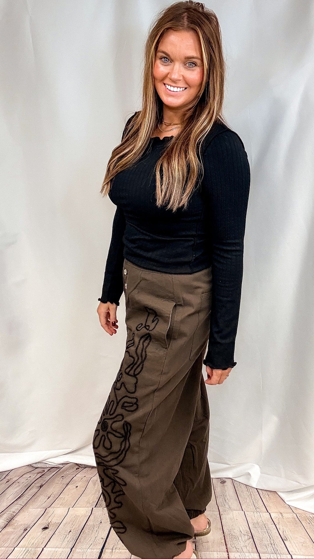 woman wearing black ribbed long sleeve top with brown pants with black embroidery on plain background