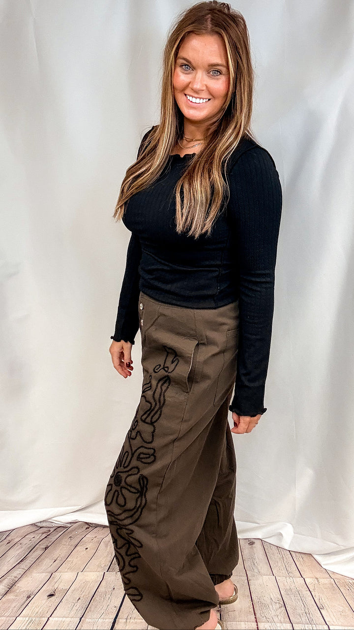 woman wearing black ribbed long sleeve top with brown pants with black embroidery on plain background