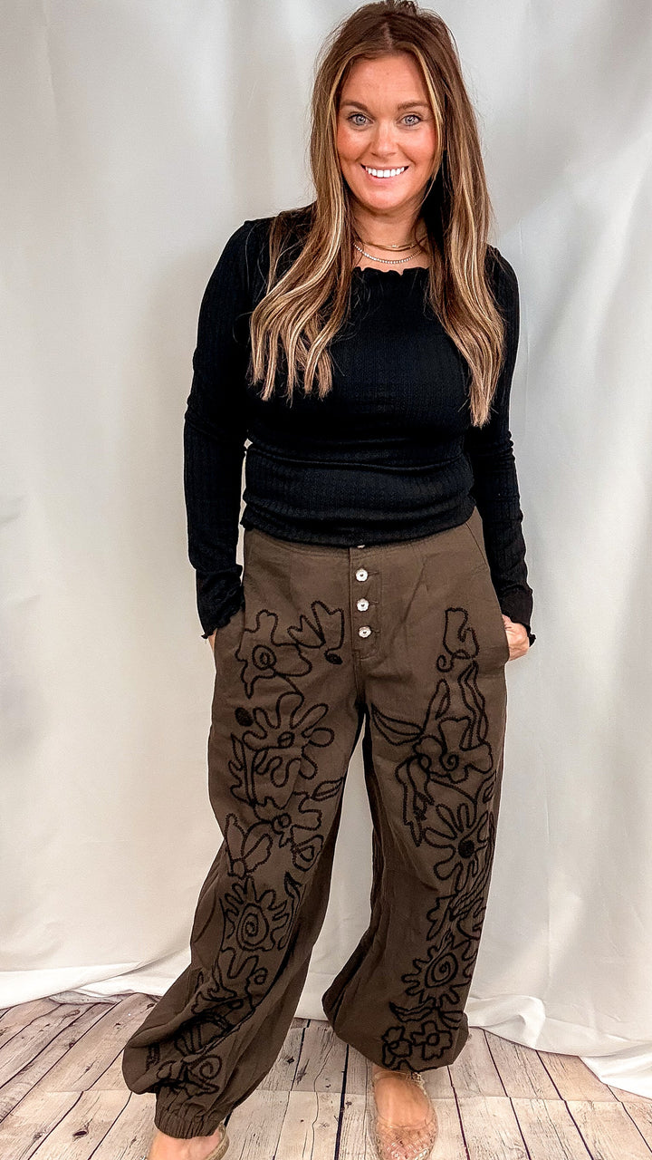 Woman wearing ribbed long sleeve black shirt with brown pants with black embroidery on front on plain background
