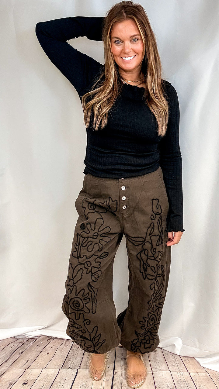 Woman wearing black long sleeve ribbed shirt with brown pants with black embroidery on front on plain background