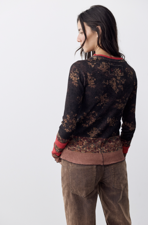 Woman shown from behind wearing black and brown printed top with red banding at neck and wrists with brown pants on white background