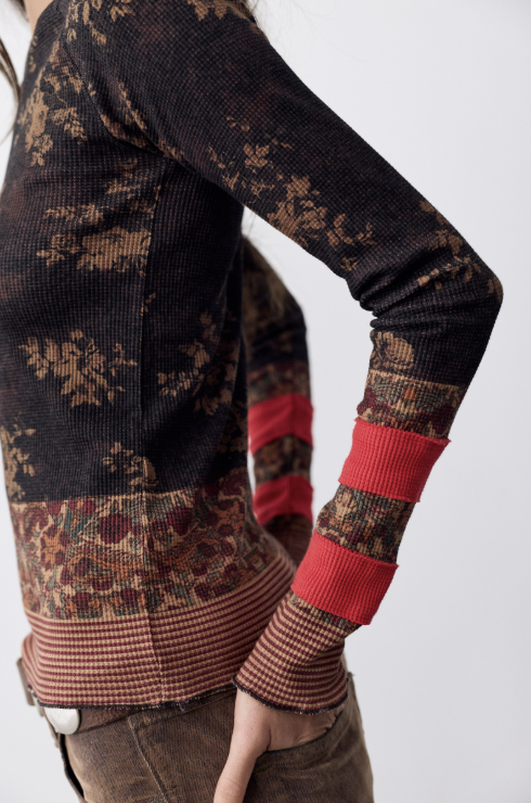 A close-up of a person wearing a long-sleeve knit sweater with a dark brown and tan floral pattern, a multicolor floral border band, and bright red ribbed cuffs.