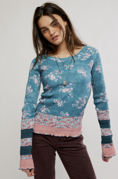 A woman modeling a long-sleeve knit sweater with a blue base, pink and white floral pattern, and striped ribbed trim, paired with dark brown pants against a light gray studio background.