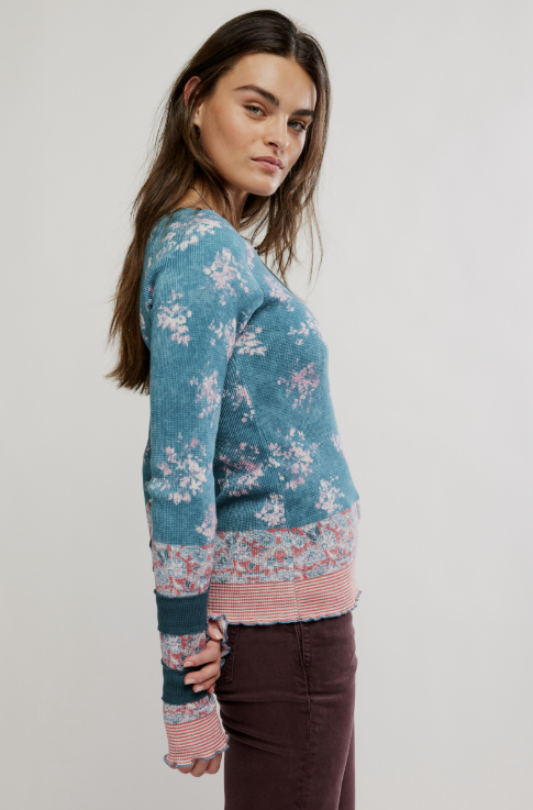 A woman wearing a teal blue knit sweater with a pink and white floral pattern, striped ribbed hem and cuffs, and dark brown pants, photographed against a plain light gray studio background.