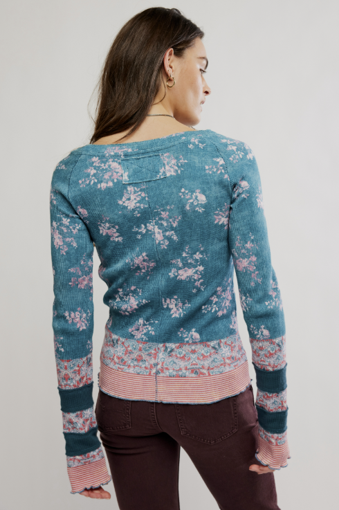 A woman shown from the back wearing a teal blue knit sweater with a pink and white floral pattern, striped trim at the hem and cuffs, and dark brown pants, against a light gray studio background.