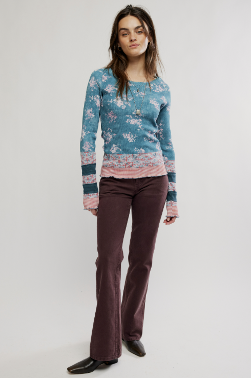 A woman modeling a long-sleeve knit sweater with a blue and pink floral pattern, paired with dark brown flared trousers and black shoes, against a plain light gray studio background.