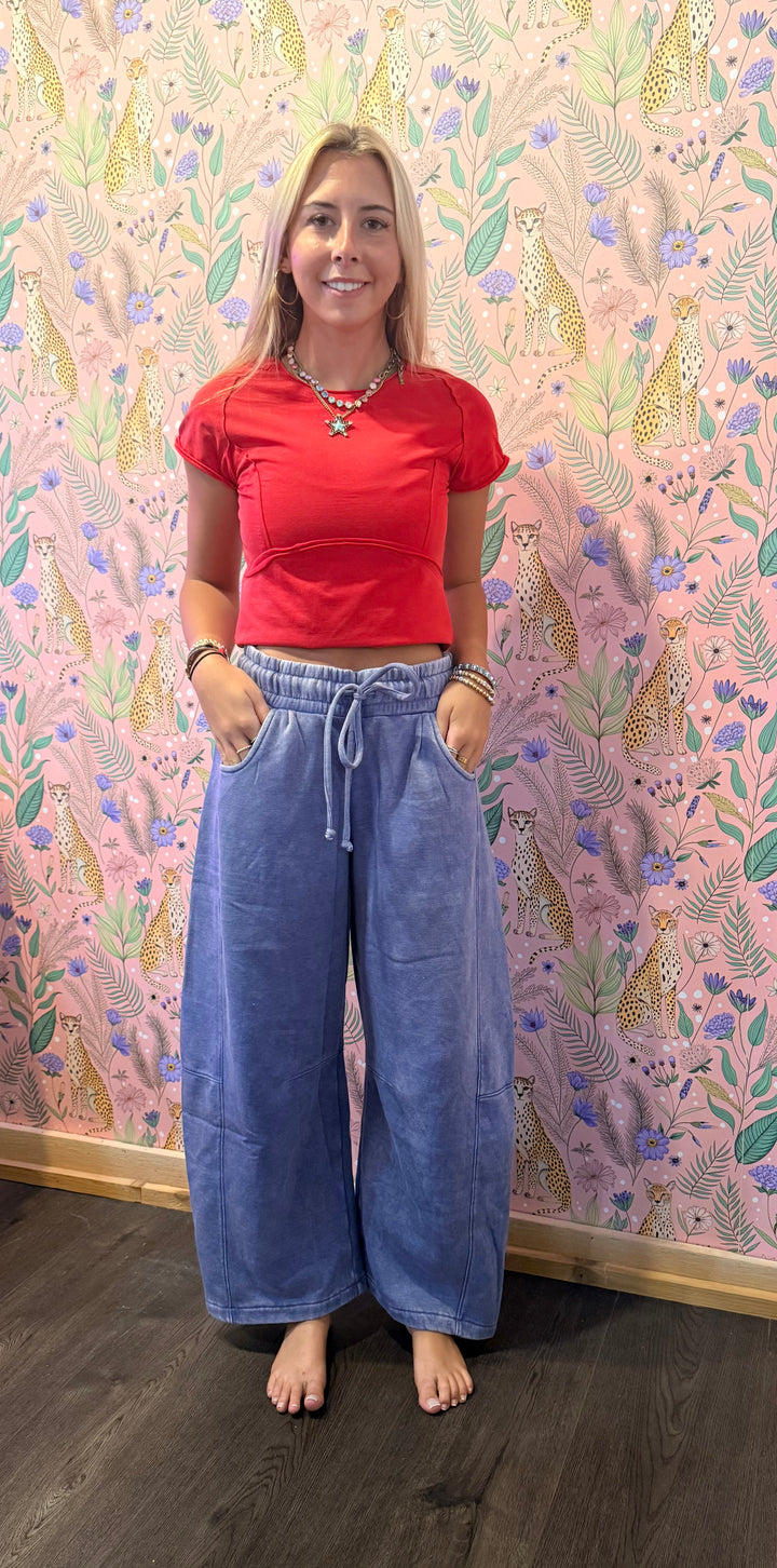 A woman stands barefoot in front of a pink floral wall tapestry, wearing a bright red short-sleeve crop top and wide-leg blue denim pants with a drawstring waist.