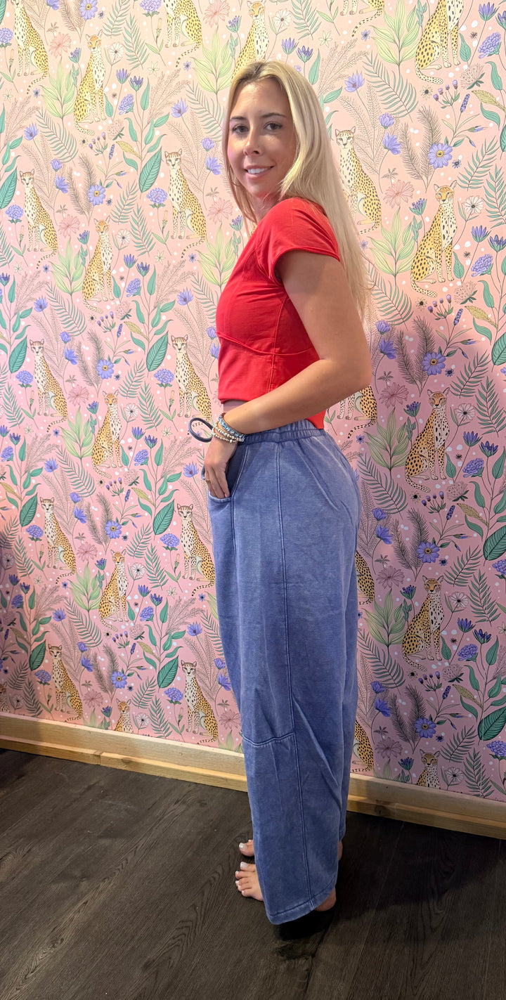 A woman stands in profile wearing a bright red short-sleeve crop top and high-waisted, wide-leg blue jeans against a pink patterned wallpaper background.