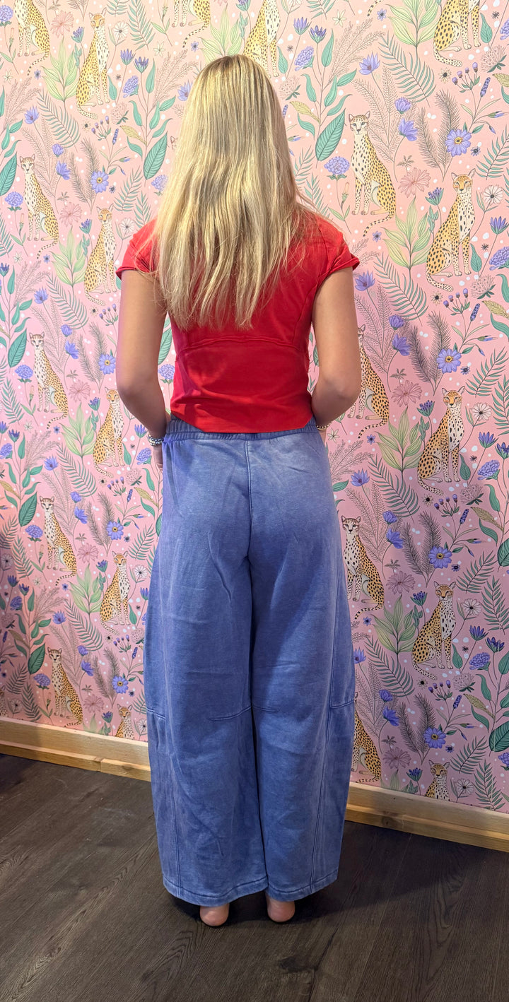 A woman with long blonde hair is shown from behind, wearing a red short-sleeve t-shirt and wide-leg blue jeans, standing in front of a pink floral wallpaper background.