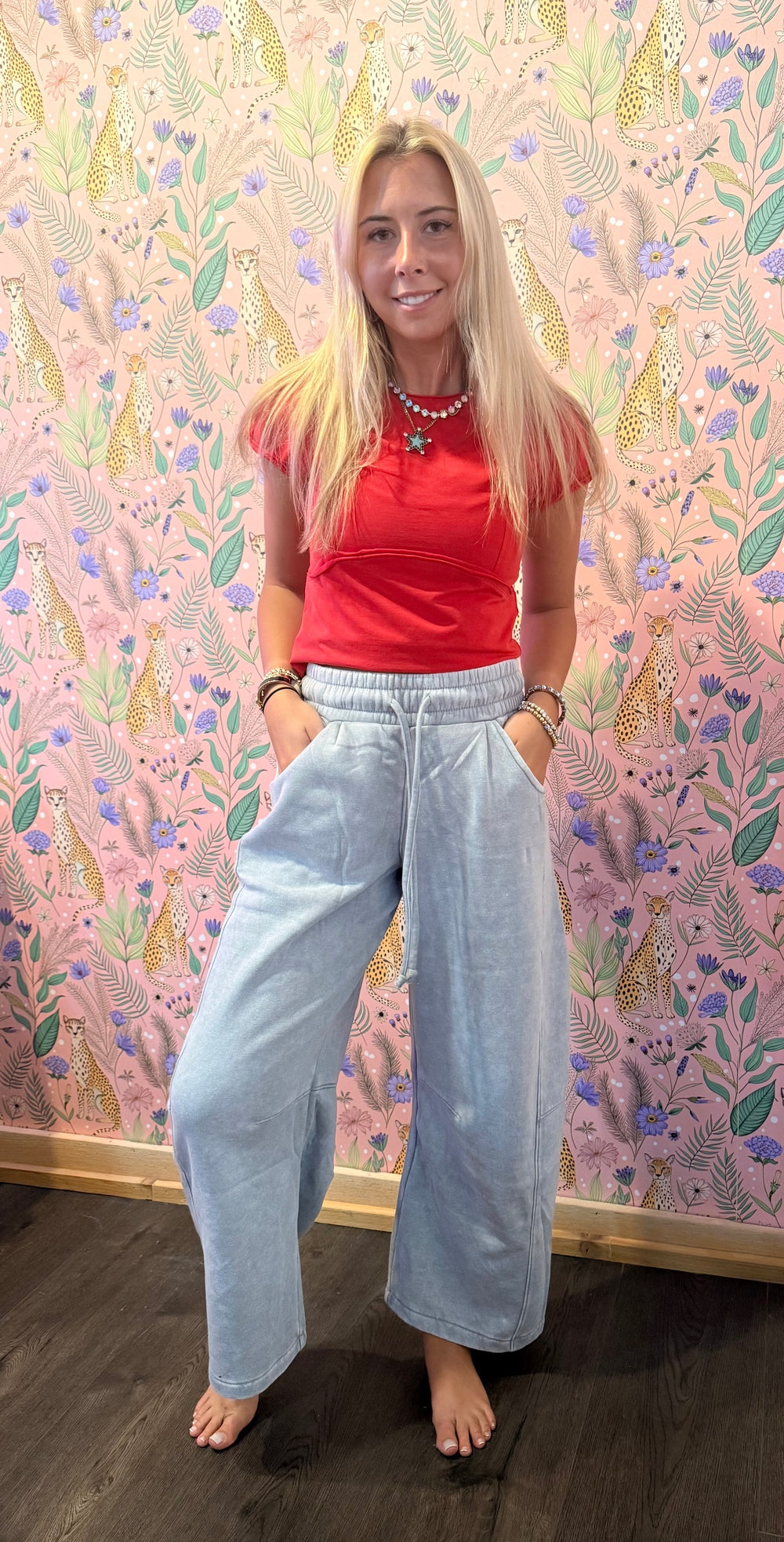 A woman stands barefoot in front of a pink floral wallpaper backdrop, wearing a red short-sleeve crop top and light-wash wide-leg jeans.