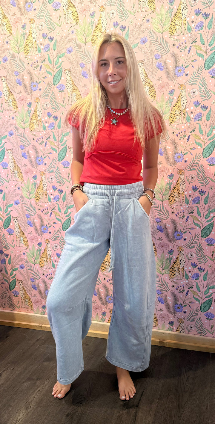 A woman stands barefoot in front of a pink floral wallpaper backdrop, wearing a red short-sleeve crop top and light-wash wide-leg jeans.
