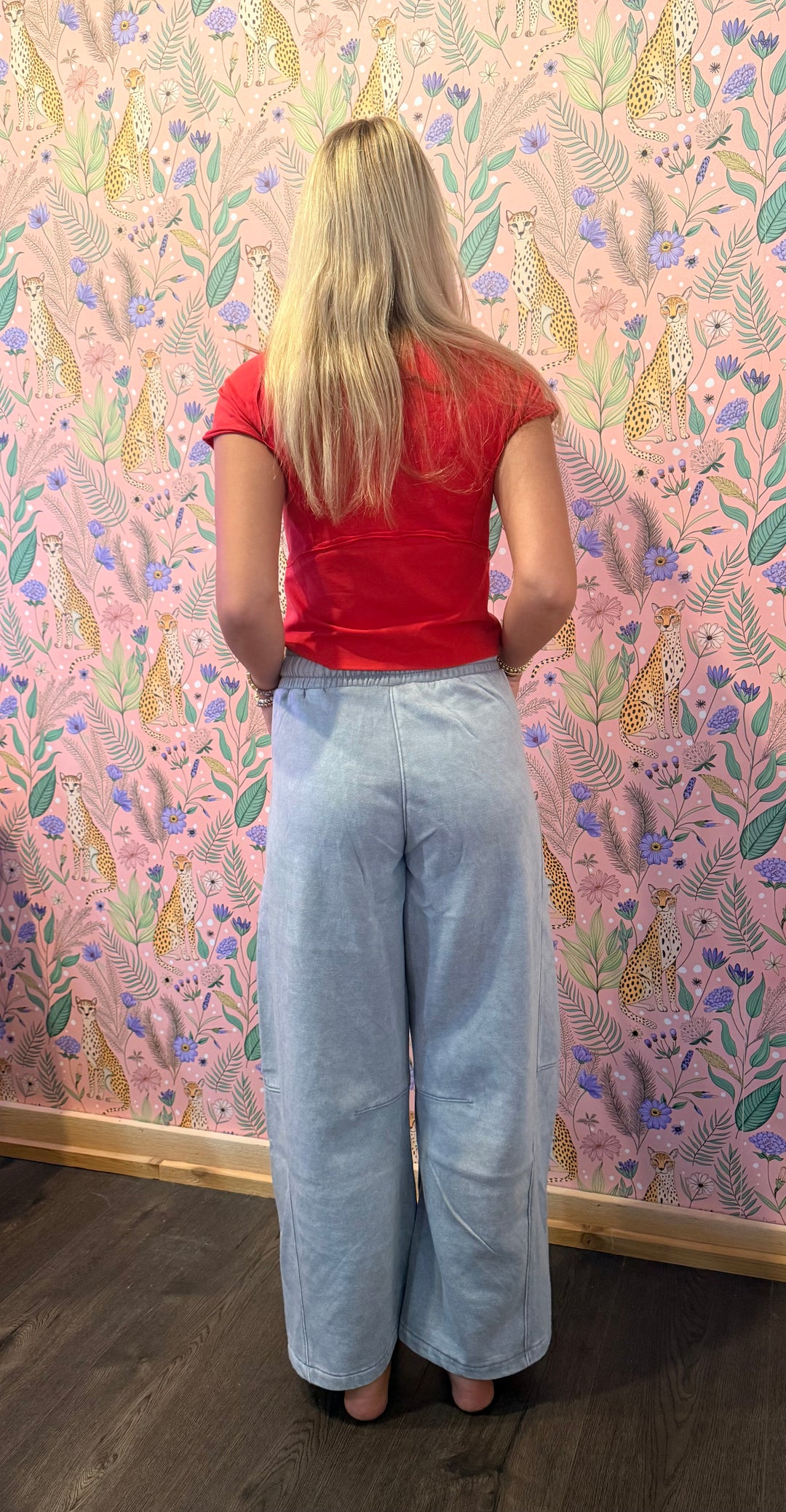 A woman viewed from behind wearing a red short-sleeve t-shirt and light-wash wide-leg denim jeans, standing in front of a pink patterned wallpaper background.