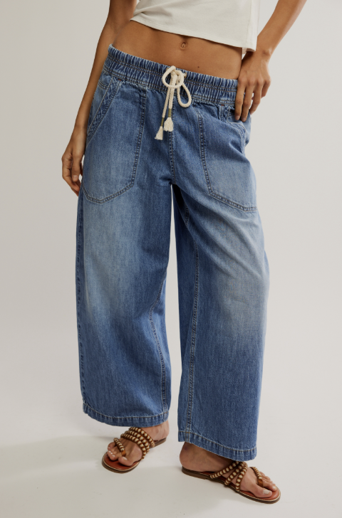 A person wearing wide-leg blue denim jeans with an elastic drawstring waistband, paired with brown studded sandals against a plain light gray studio background.