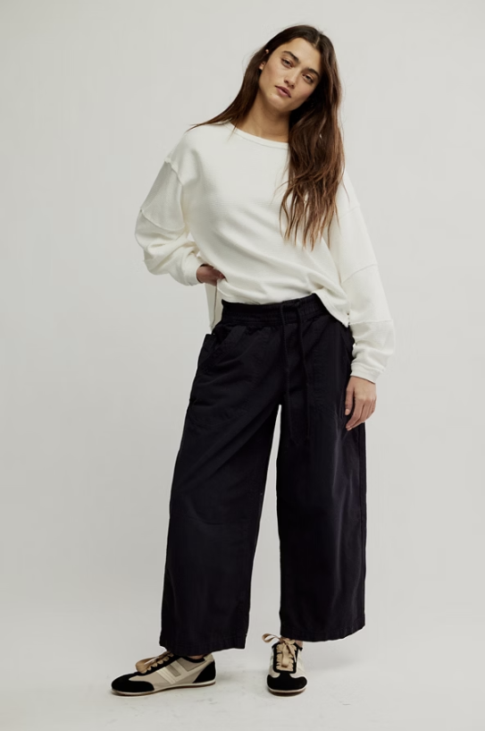A woman modeling a white long-sleeve crewneck sweatshirt and black wide-leg trousers, paired with black and beige sneakers, against a plain light gray studio background.