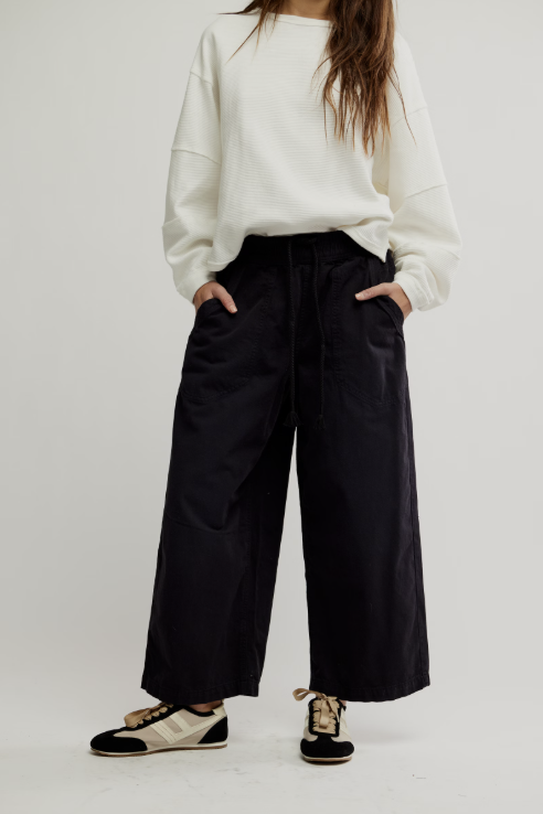 A model wearing a white crewneck sweatshirt, black wide-leg cropped pants, and black-and-white sneakers, standing against a light gray studio background.