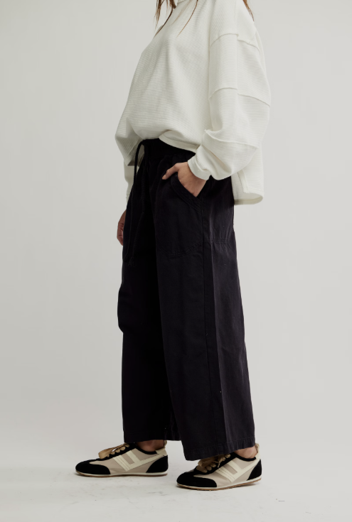 A model is shown from the chest down wearing black wide-leg trousers with side pockets, paired with a white long-sleeve top and black-and-white sneakers, against a plain light gray studio background.