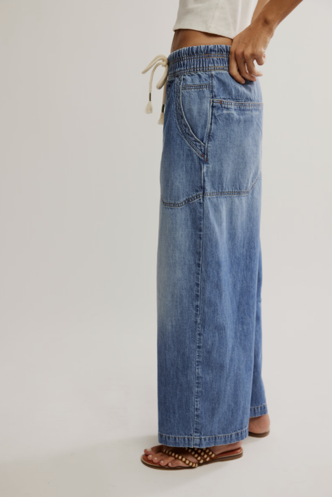 A person wearing a pair of wide-leg blue denim jeans with an elastic waistband and cream drawstring, paired with brown strappy sandals against a plain light gray studio background.