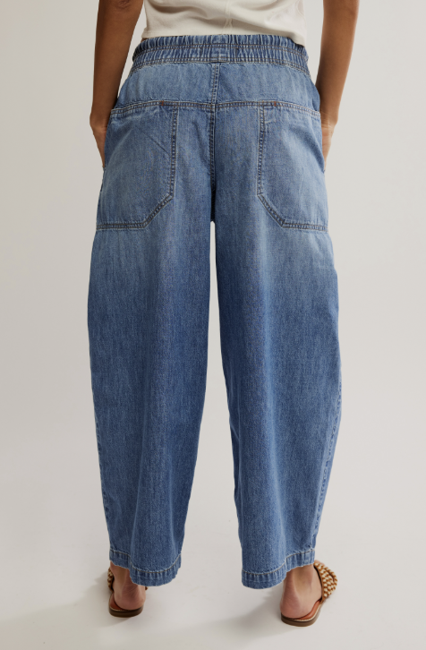 Rear view of a person wearing wide-leg blue denim jeans with an elastic waistband and back patch pockets, paired with a white top and brown heeled sandals against a light gray studio background.